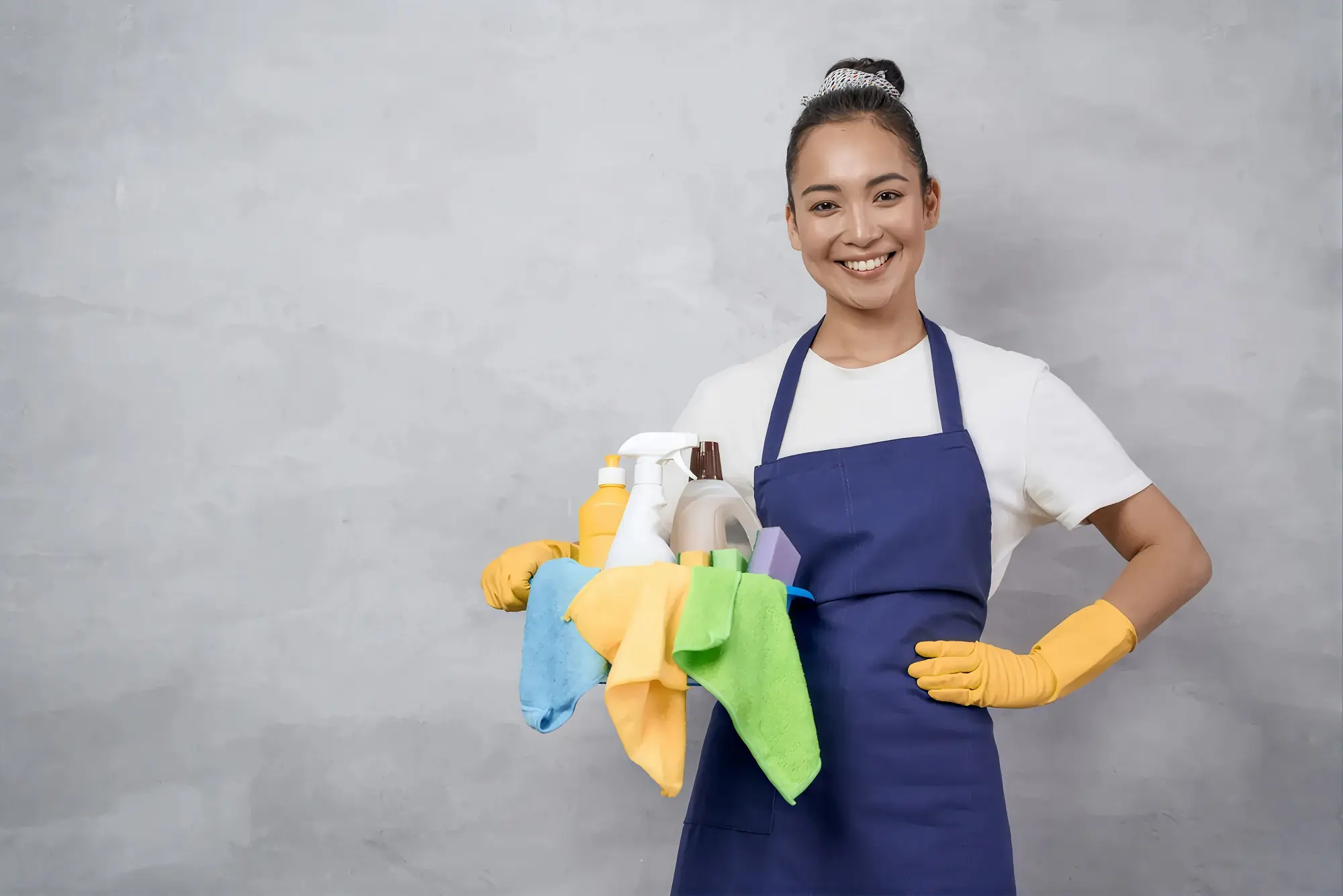 Colaboradora sorridente de avental azul e luvas, segurando um balde com produtos de limpeza